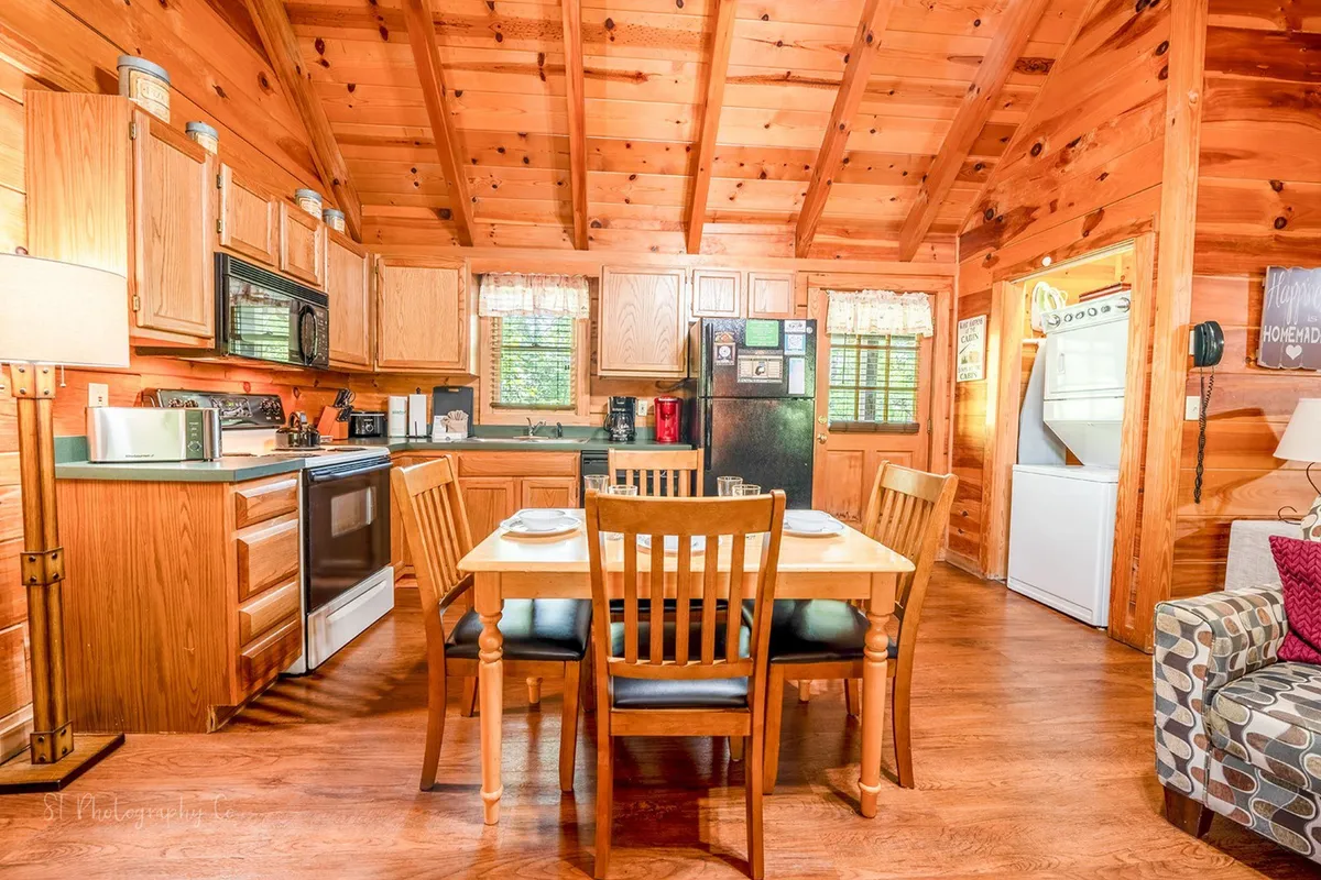 A welcoming open-concept kitchen and dining area featuring wooden kitchen cabinets, a well-lit dining table set for four, and a comfortable seating area nearby. The kitchen is equipped with modern appliances and ample counter space for meal preparation. Warm lighting and rustic wood tones create a cozy, inviting ambiance, perfect for cooking and gathering with family.