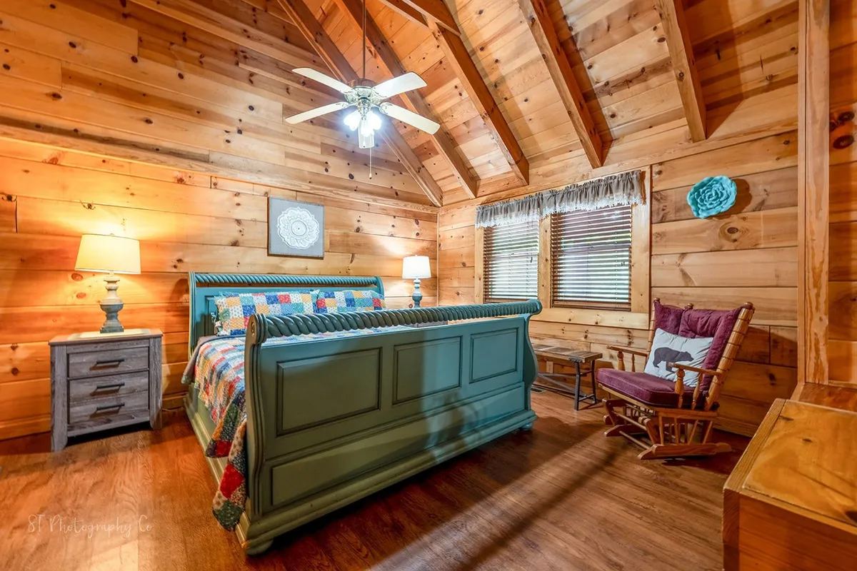 A cozy bedroom scene with a colorful quilt, blue bed frame, wooden ceiling, nightstands with lamps, and a rocking chair. The room feels welcoming and homey, suggesting a restful place to recharge after outdoor activities.