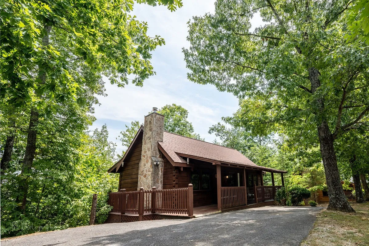 This image captures the log cabin's exterior, showcasing the rustic structure, stone chimney, and surrounding woodland. The front porch invites relaxation while the backdrop of trees hints at the secluded mountain setting. The overall atmosphere is peaceful and intimate, aligning with the section's themes of romance and natural beauty.