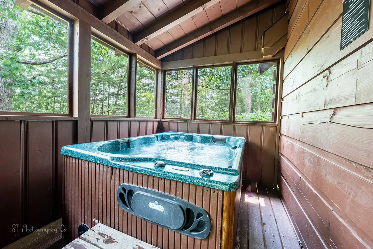 This interior image highlights a screened-in porch with a private hot tub, wooden walls, and large windows overlooking woodland scenery. The tranquil ambiance is perfect for soaking, stargazing, or enjoying coffee while immersed in nature.