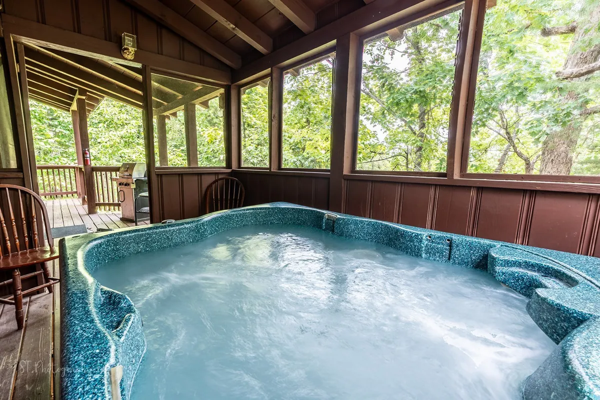 A cozy hot tub sits surrounded by wooden walls and screened windows, allowing natural light and nature views to filter in. The tranquil setting is further enhanced by the sense of privacy provided by the screened-in porch, with plenty of space for relaxation and conversation. The view extends to the surrounding trees, immersing guests in the cabin’s natural environment while enjoying the bubbling hot tub.