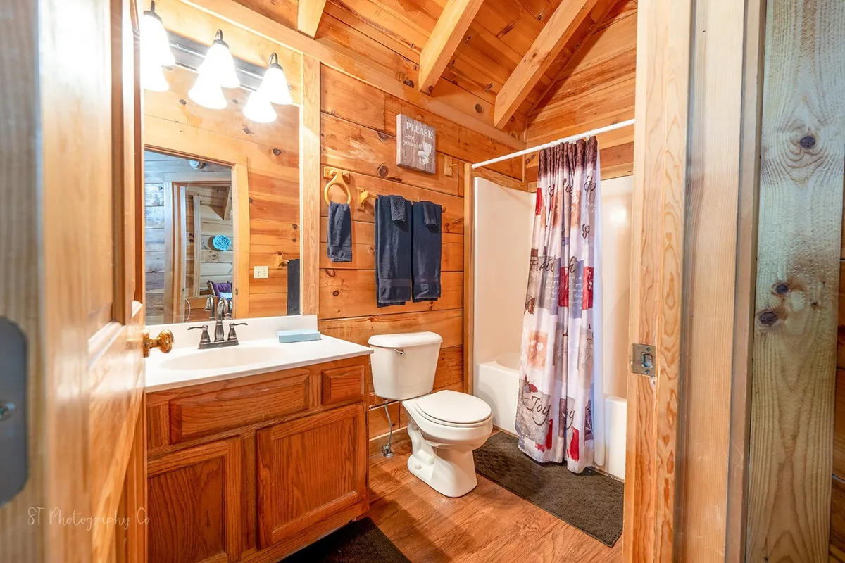 A shared bathroom with wooden walls, a vanity with mirror, toilet, and shower curtain. The warm lighting and inviting finishes convey both comfort and privacy, with ample space for personal grooming and relaxation.