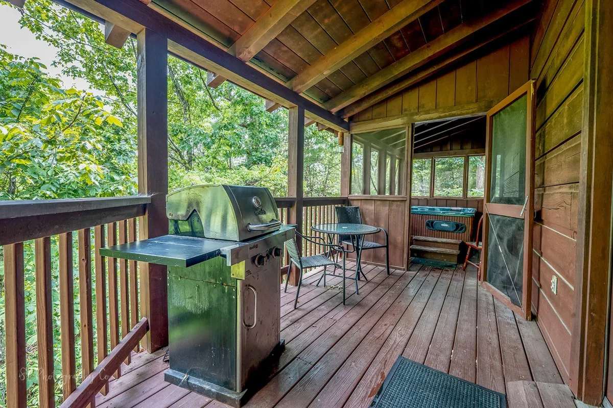 A gas grill stands ready on a spacious deck, accompanied by a table and chairs for al fresco dining. Surrounded by a wooden railing and the lush greenery of the Smoky Mountains, this outdoor space is designed for grilling, socializing, and savoring meals in the fresh mountain air.