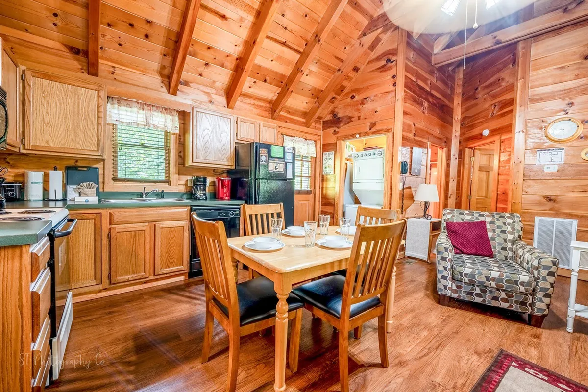 A bright, open-plan kitchen and dining space accented by a wooden ceiling, a prominent dining table for four, and a view of the kitchen area with appliances. An armchair in the corner hints at the transition from dining to relaxation. The scene feels intimate yet spacious, centered around togetherness and comfort.