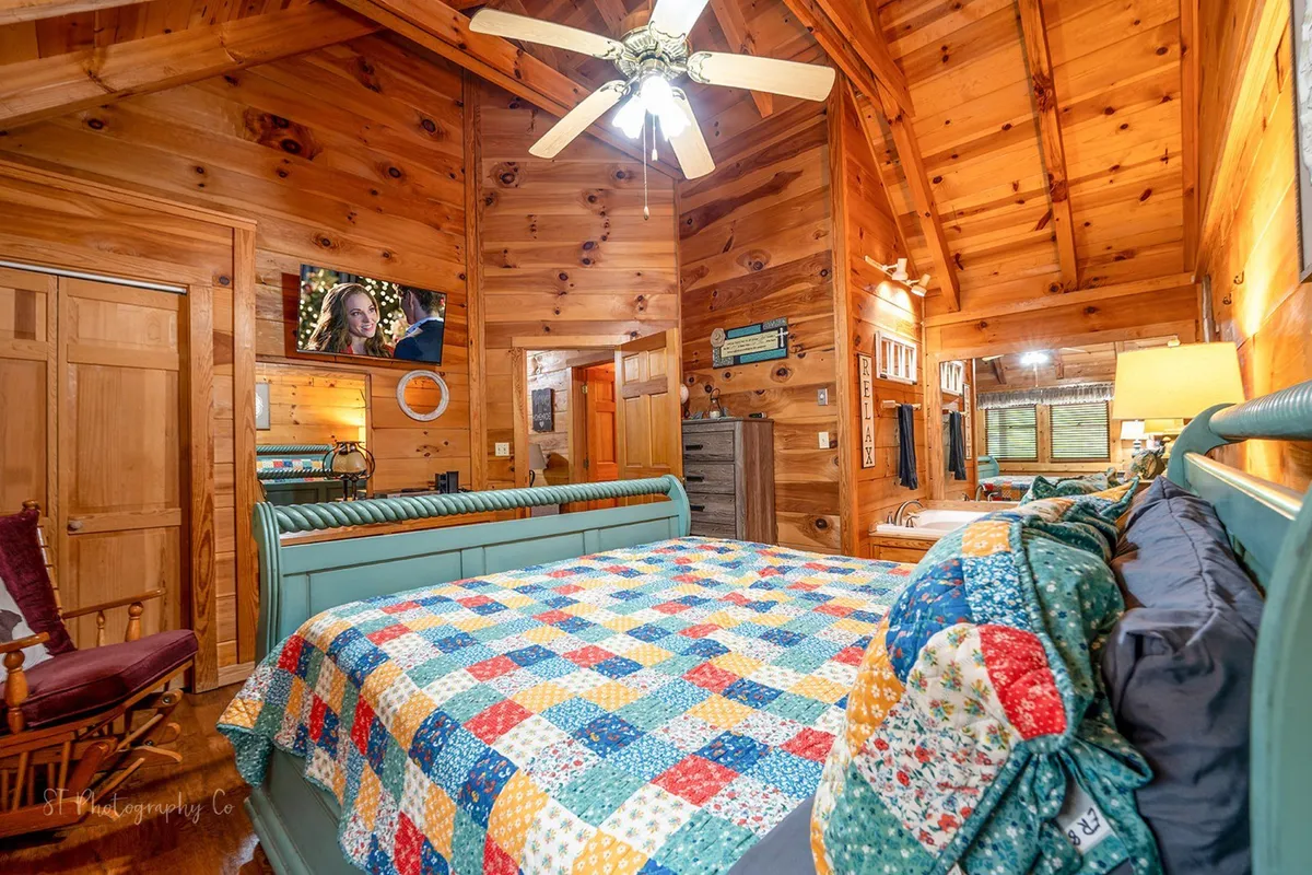 A cozy master bedroom featuring a king-sized bed adorned with a colorful quilt, wood-paneled walls, a ceiling fan, and a television mounted on the wall. The warm lighting and inviting textures emphasize a restful and tranquil atmosphere, ideal for unwinding after a long day.